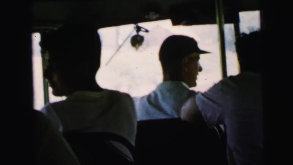 Silhouetted bus passengers in a dark vehicle interior, one in a baseball cap in profile, from an 8mm home movie circa 1964.