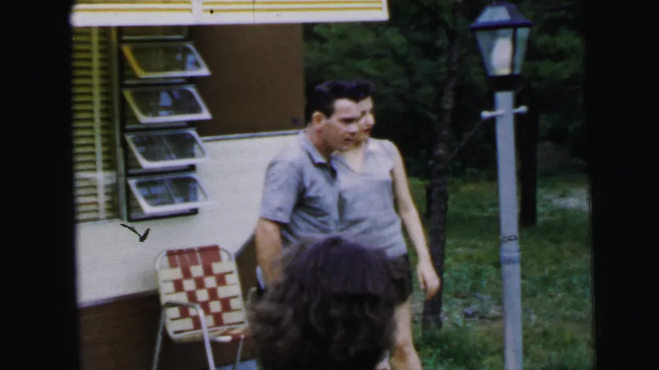 A man in a casual short-sleeved shirt stands before a 1960s travel trailer with louvered side panels; a checkered vinyl lawn chair and lamp post appear in this 1964 8mm home movie.