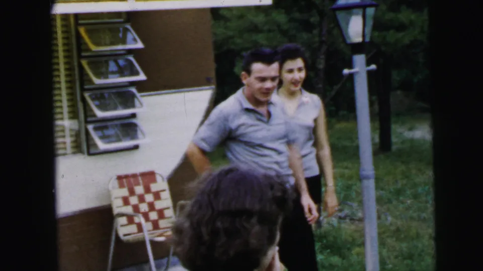 A man and woman in casual summer attire stand near a period travel trailer; a woven lawn chair and lamp post frame the scene in this 1964 8mm home movie.