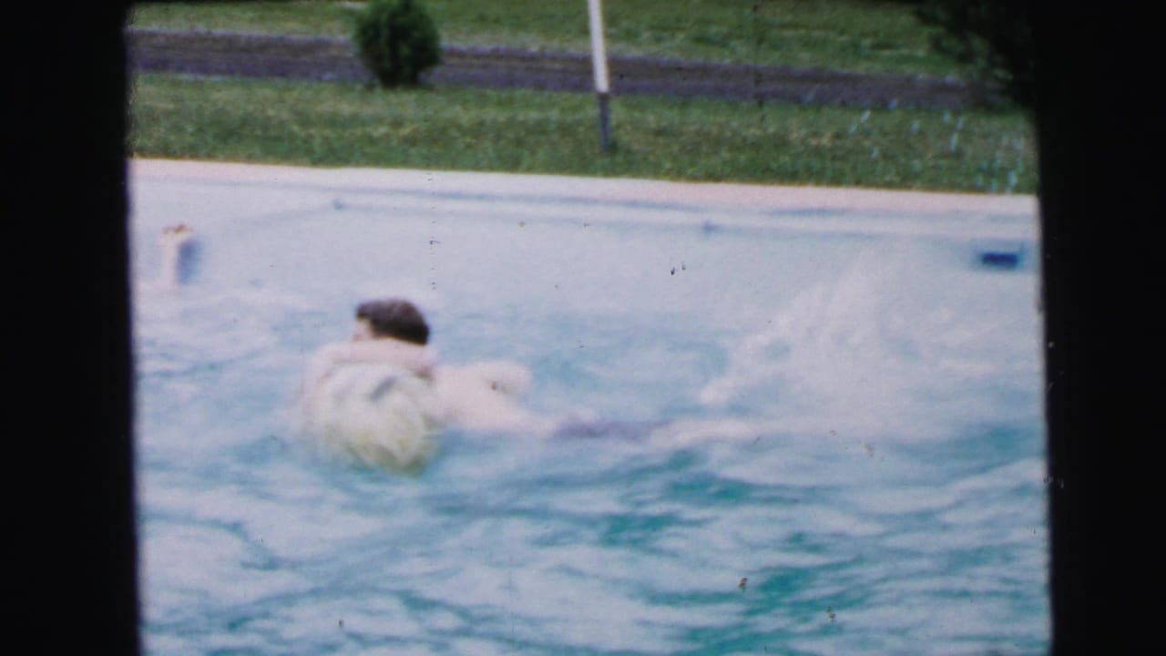 Swimmer seen from behind reaches toward a large pale beach ball floating in blue-green pool water.