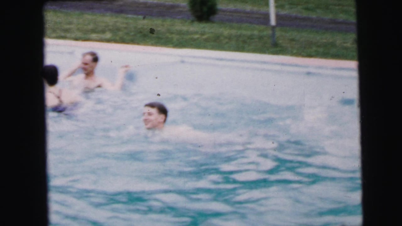 Two men in a swimming pool; the younger man laughs openly toward the camera while an older man is visible behind him.