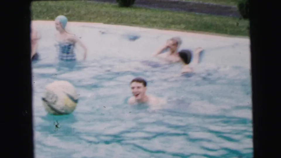 Several adults play in a sunlit backyard pool circa 1964; a woman in a gingham swimsuit and light-blue rubber swim cap stands near a large beach ball as a young man laughs at center.