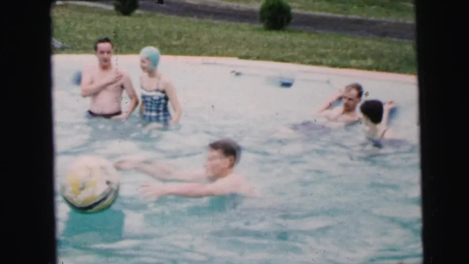 Young man catches a large ball at the pool edge while a woman in a gingham swimsuit and light-blue swim cap and other adults look on; 8mm home movie, possibly Camden, New Jersey, circa 1964.