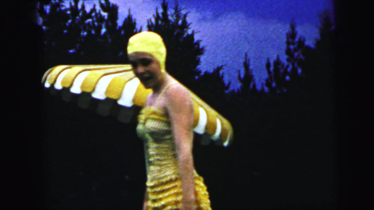 Woman in a 1960s-style yellow rubber swim cap and tiered knitted swimsuit poses with a striped patio umbrella poolside, from 1964 home-movie footage from Camden, New Jersey.