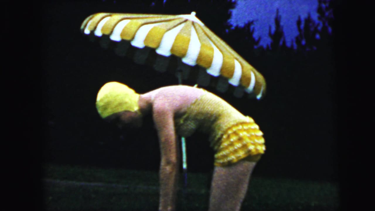 Woman in a 1960s yellow knitted swimsuit bends forward preparing to dive, with a striped patio umbrella above, in this 1964 home-movie frame from Camden, New Jersey.