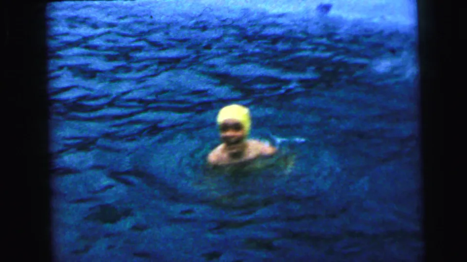 Woman in yellow rubber swim cap surfaces and looks toward camera in a deep-blue backyard pool, from a 1964 home-movie sequence attributed to Camden, New Jersey.