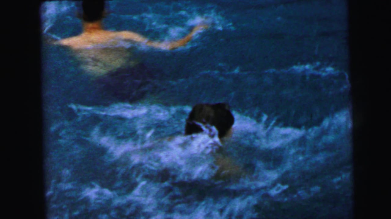 Young man partially submerged in a swimming pool, water churning around him; second shirtless figure visible at top edge of frame.
