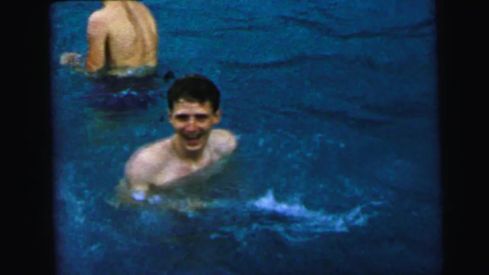 Young dark-haired man smiling directly at the camera while standing in a swimming pool; a second shirtless person is visible behind him; 8mm home movie footage, Camden NJ, circa 1964.