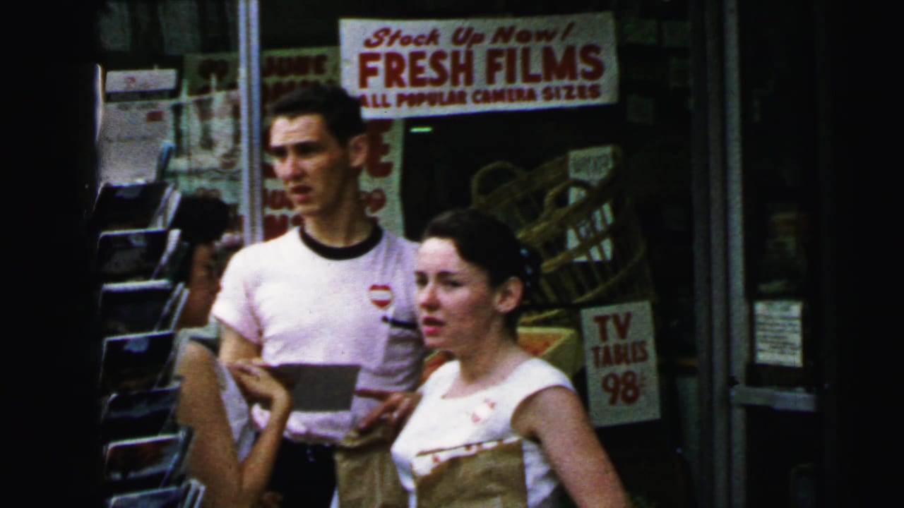 Young couple standing before a 'FRESH FILMS' camera-supplies storefront; man in white ringer t-shirt, woman in white sleeveless top carrying a brown bag.