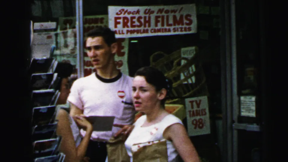 Young couple on a Camden, New Jersey sidewalk in 1964, pausing before a storefront advertising 'FRESH FILMS' camera supplies and 'TV TABLES 98¢'.