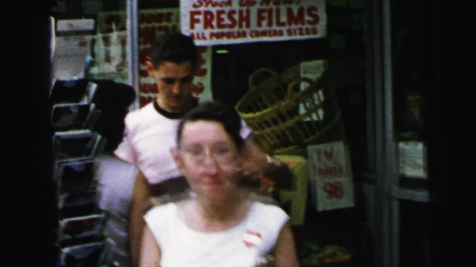 Young couple outside a Camden, NJ camera-supplies shop in 1964; man smiling, woman turning, retail signage prominent in background.