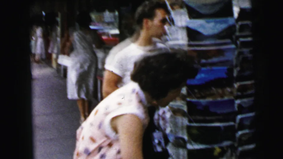 Woman browsing a sidewalk merchandise rack in 1964, likely Camden, NJ; a passerby in a floral dress visible to her left.
