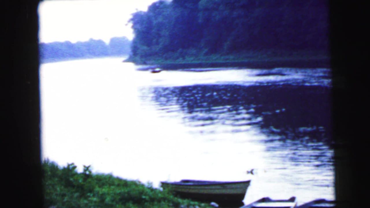 Small rowboat resting on grassy riverbank, calm water stretching to a tree-lined far shore, 8mm film.