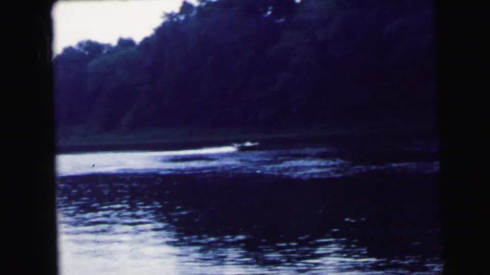Motorboat with visible white wake crossing a wide river, dark dense tree line on far shore; 8mm home movie attributed to Camden, New Jersey, 1964.
