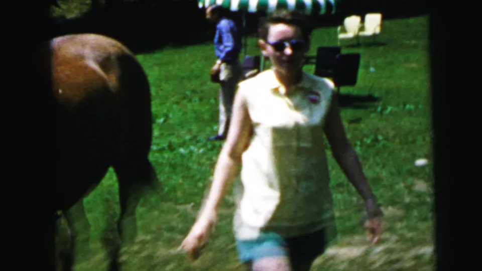 Woman with sunglasses turns toward camera on a sunny lawn beside a brown horse, striped awning and other visitors visible behind, 8mm home movie, circa 1964.