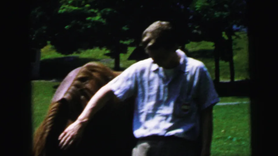 Man in light blue shirt strokes a brown horse on a sunlit lawn, trees visible in background, 8mm home movie, Camden, New Jersey area, circa 1964.