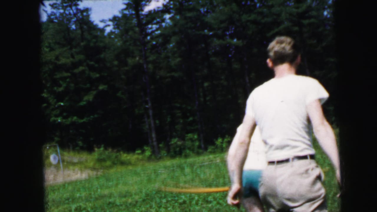 Man in white t-shirt seen from behind on a grass field, with an evergreen tree line and a distant archery target visible.