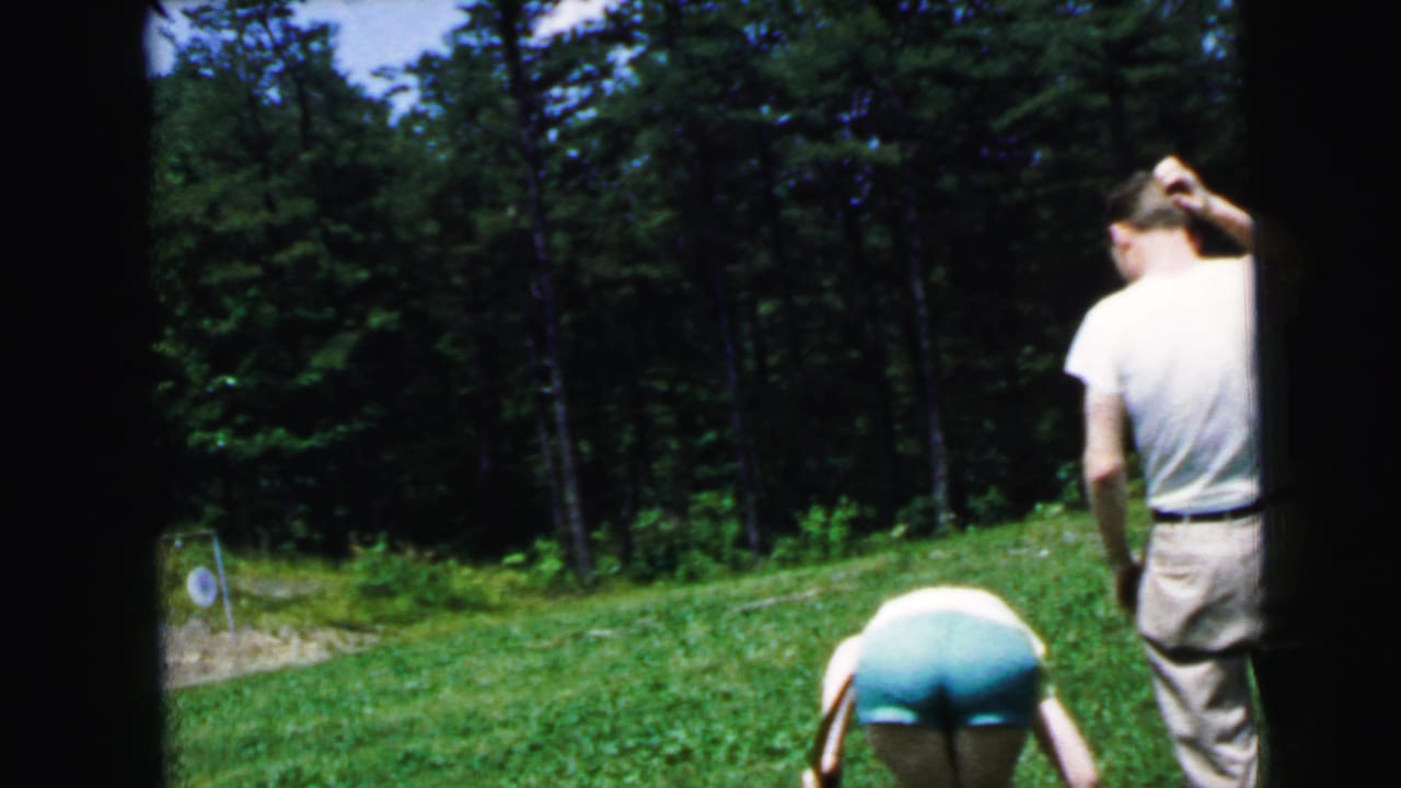Man in white t-shirt standing with hand raised to head; woman in teal shorts bending toward ground on a grass archery range.