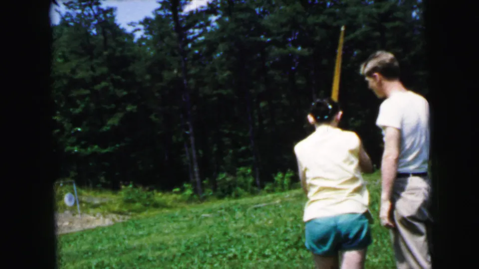 Woman holding an oversized wooden recurve bow and man standing beside her, both viewed from behind on an outdoor archery range circa 1964, dense evergreen trees and hillside target behind.