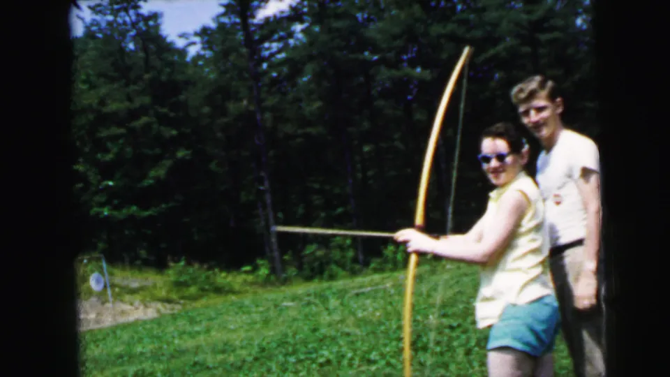 Woman in yellow sleeveless top, teal shorts, and cat-eye sunglasses holds a large wooden recurve bow posing with a man in a white t-shirt on a grass archery range, circa 1964.