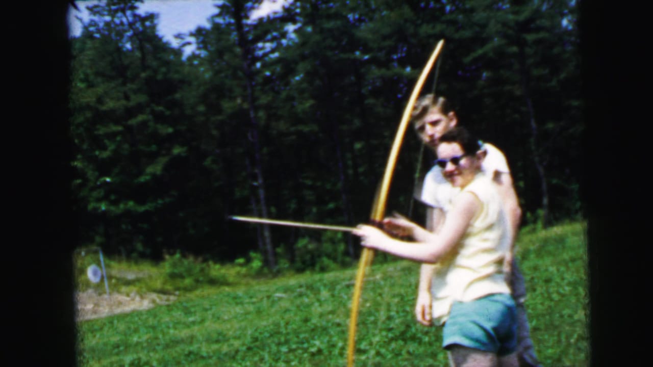 Woman in yellow sleeveless top and blue shorts drawing a longbow, guided by a man in a white shirt, on a grassy slope with dense trees behind.