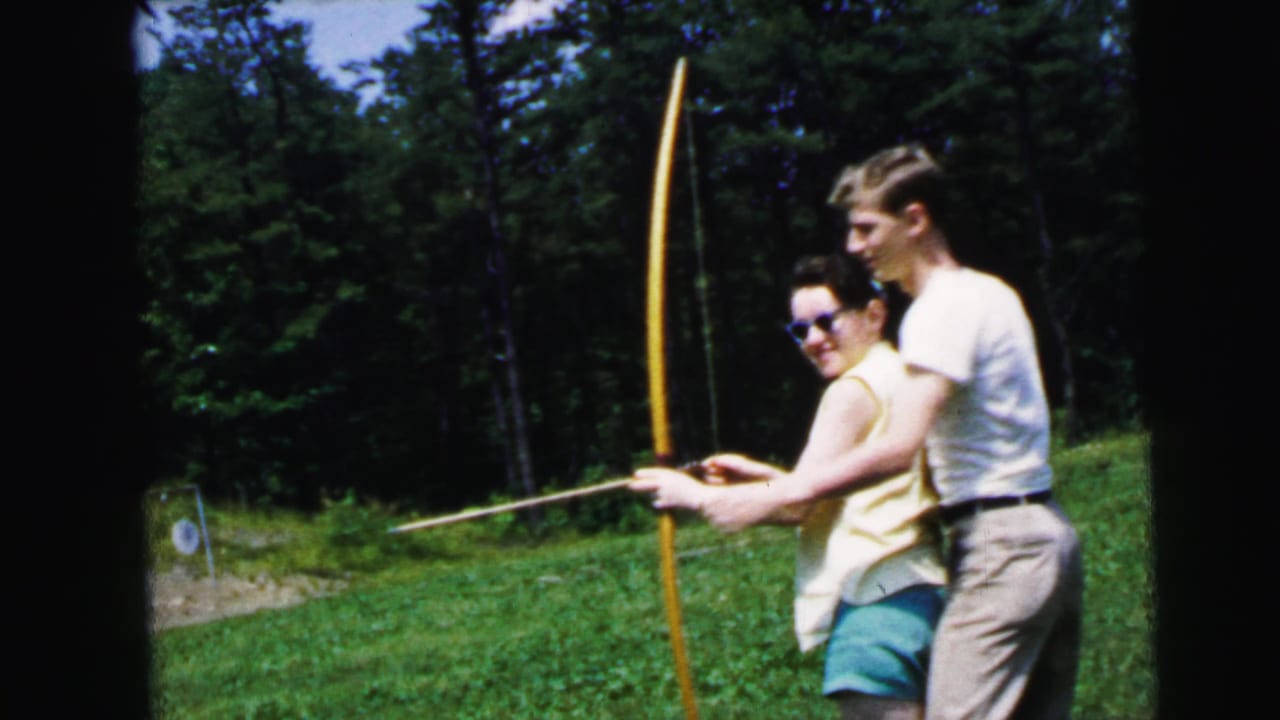 Woman in yellow top and blue shorts at full draw on a yellow longbow while a man in a white shirt guides her stance; 8mm film, New Jersey, 1964.