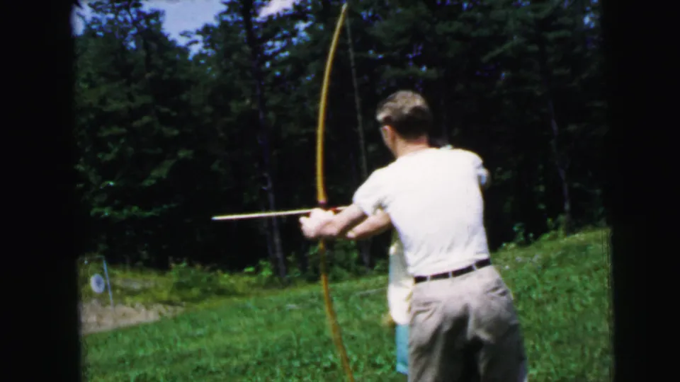 Man in white short-sleeve shirt and khaki trousers seen from behind drawing a yellow longbow; 8mm home movie, New Jersey, 1964.