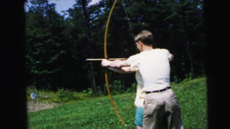 Man at full draw on a yellow longbow aiming at a circular archery target on a green slope backed by dense trees; 8mm home movie, New Jersey, 1964.