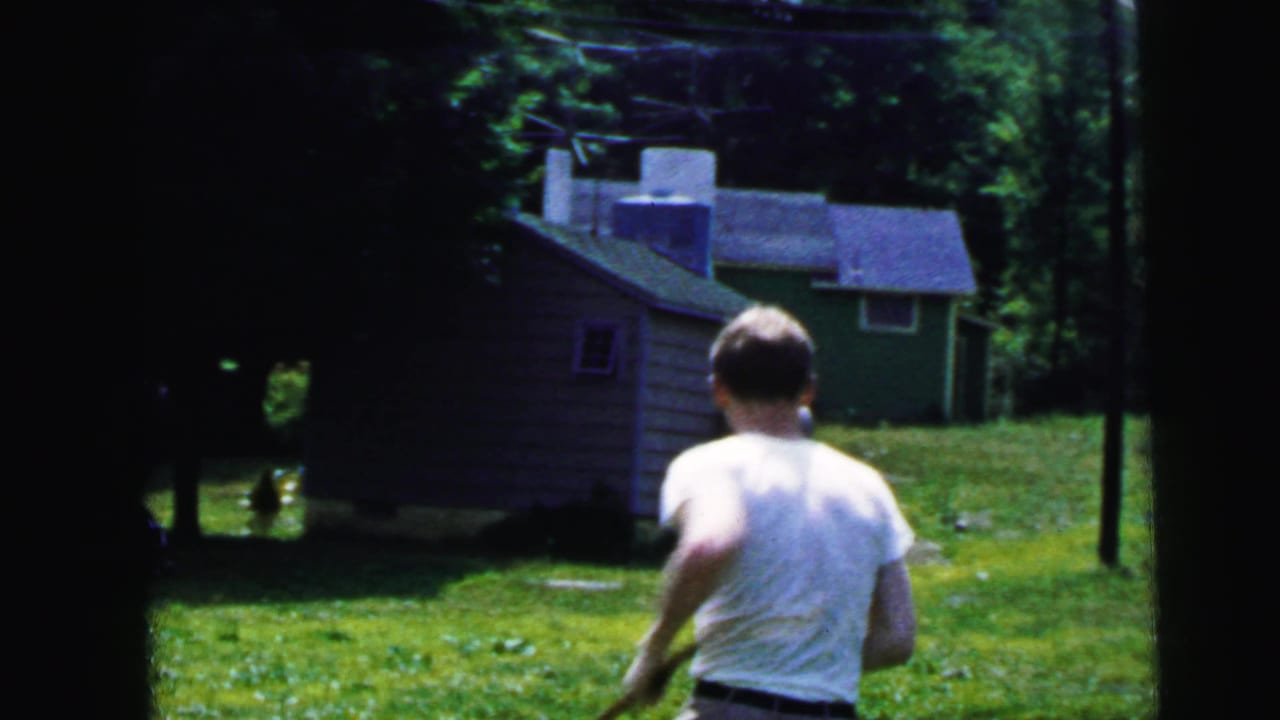 Man in a white shirt runs across a grassy Camden yard in 1964, moving toward small outbuildings under trees.