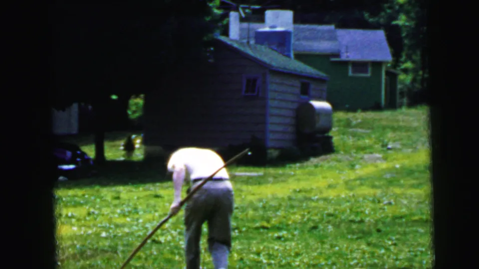 A person bends on a green lawn near a small shed in 1964 Camden home-movie footage, with a long stick visible across the frame.