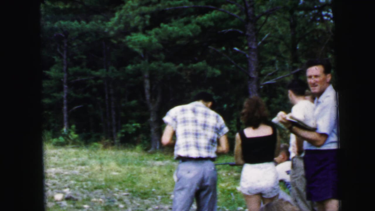 Group of adults in wooded campground clearing; man with clipboard faces two others in summer clothing.