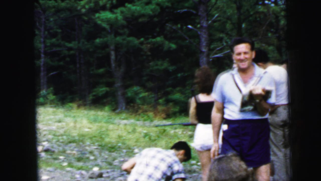 Man smiling at camera during campground game activity circa 1964; holding papers, rocky terrain and pine forest background visible.