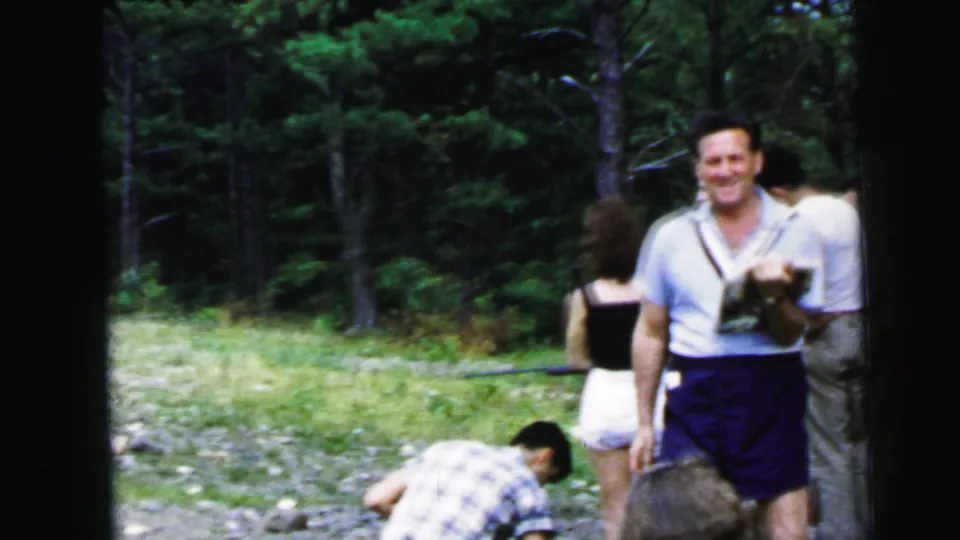 Man smiling at camera during campground game activity circa 1964; holding papers, rocky terrain and pine forest background visible.