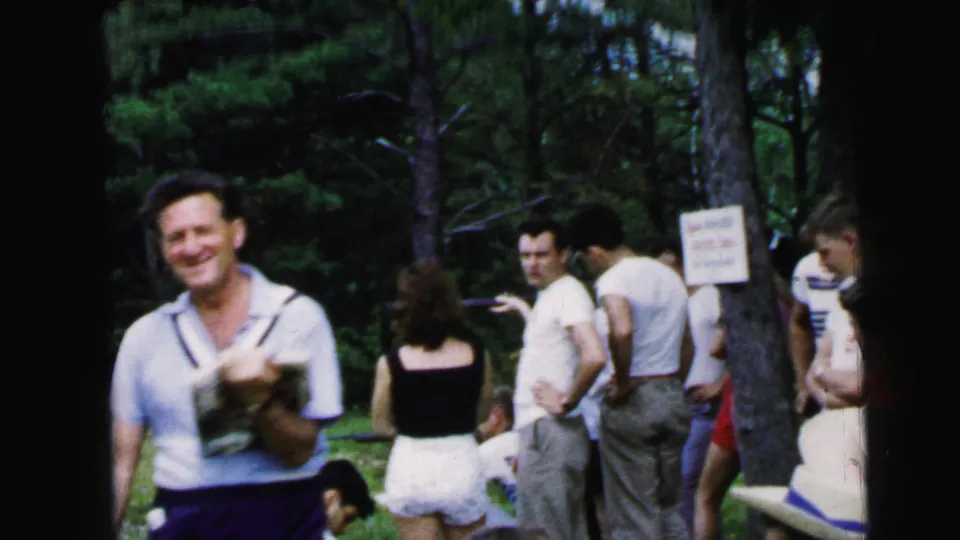 Group of adults at campground game station circa 1964; posted sign near tree trunk, pine trunks, man in light blue shirt at left.