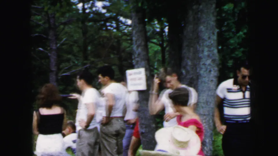 Group of adults at campground game activity circa 1964; woman in wide-brimmed straw hat, man in striped shirt, pine trees and posted sign.