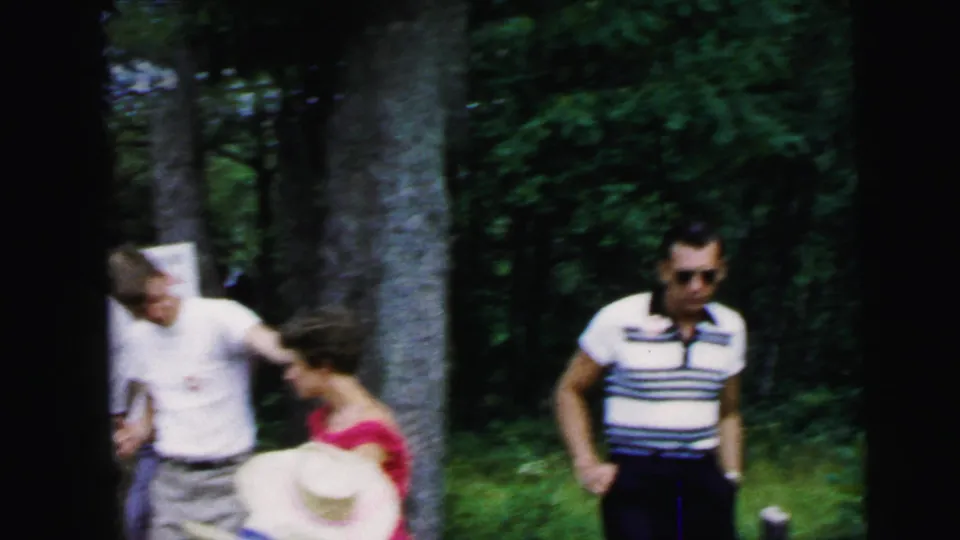 1964 8mm home movie: adults gathered in a wooded clearing near Camden, NJ; man in striped polo and sunglasses, woman in red dress, straw-hatted figure.