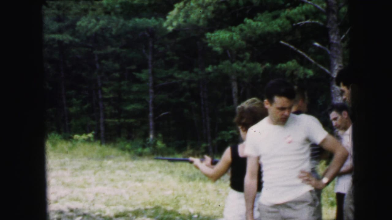 Man in white t-shirt stands in foreground while a woman aims a long rifle; other adults watch in a sunny wooded clearing.