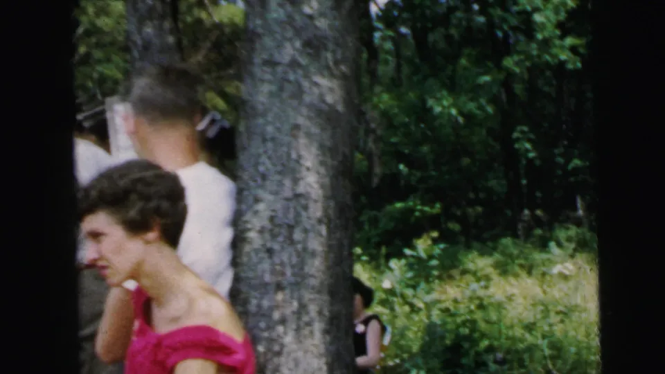 Close-up frame from a 1964 8mm home movie: woman in bright pink top facing sideways in a wooded setting consistent with the Camden, New Jersey location.