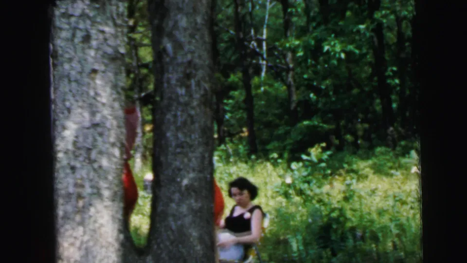 8mm home movie frame, 1964: two large tree trunks in wooded clearing near Camden, NJ; seated woman in dark clothing at right, red-dressed figure visible between trees.
