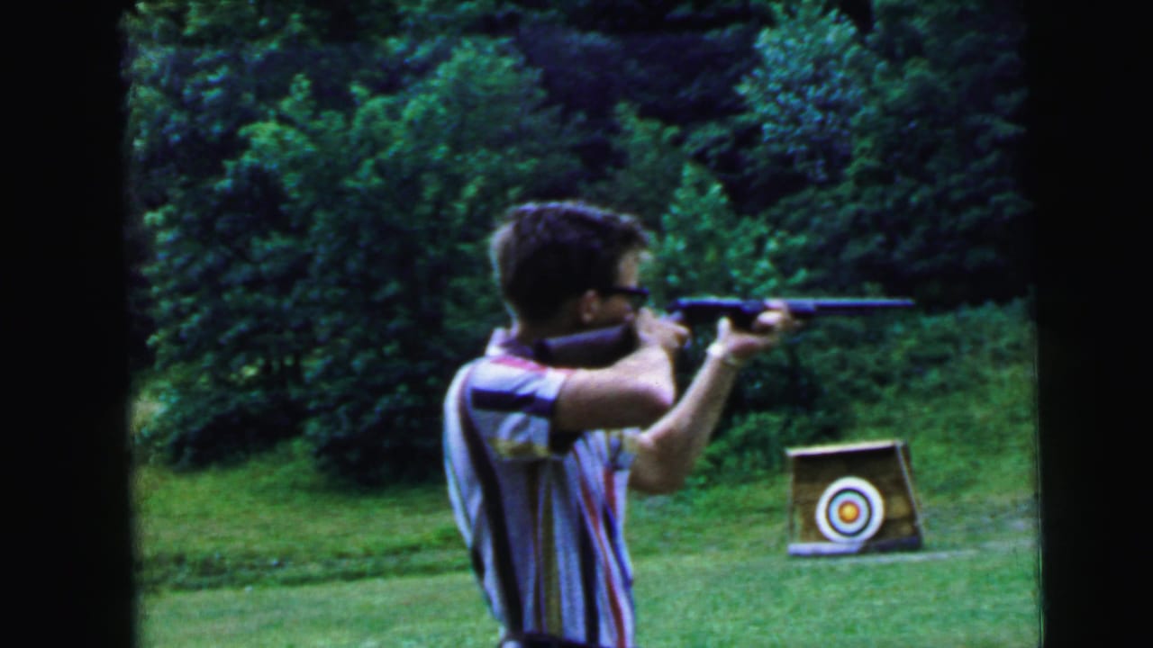 1964 8mm home movie frame of a young man in a striped shirt and dark-rimmed glasses taking aim with a rifle at a bullseye target on a stand, with a second target and dense summer foliage in the background.