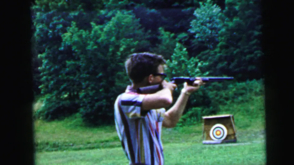 Mid-sequence 8mm home movie frame of a young man with dark-rimmed glasses and a striped shirt shouldering a rifle aimed at a circular bullseye target on a wooden stand, summer woodland backdrop.