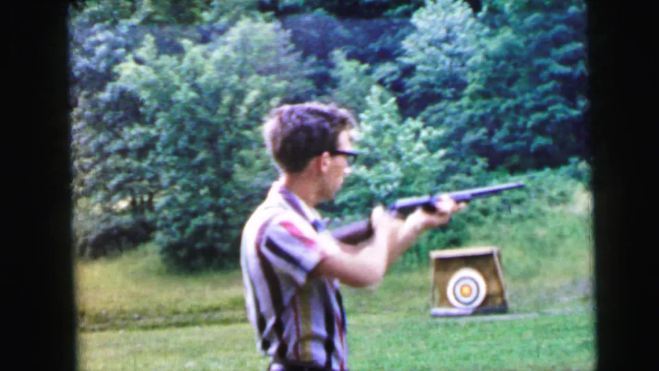 Final 8mm frame of a young man in a striped shirt and dark-rimmed glasses aiming a rifle at a bullseye target on a stand, captured in warm natural light against a lush summer green backdrop.
