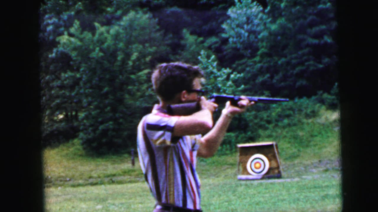 Young man in striped shirt and glasses aiming a rifle at a bullseye target in an outdoor grassy field, 8mm home movie.