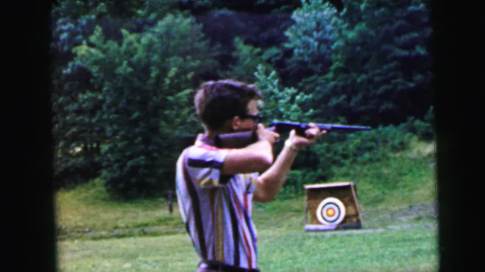 Young man in striped shirt and glasses takes aim with a rifle at a circular bullseye target in a grassy outdoor field; Camden NJ, 1964 8mm home movie.