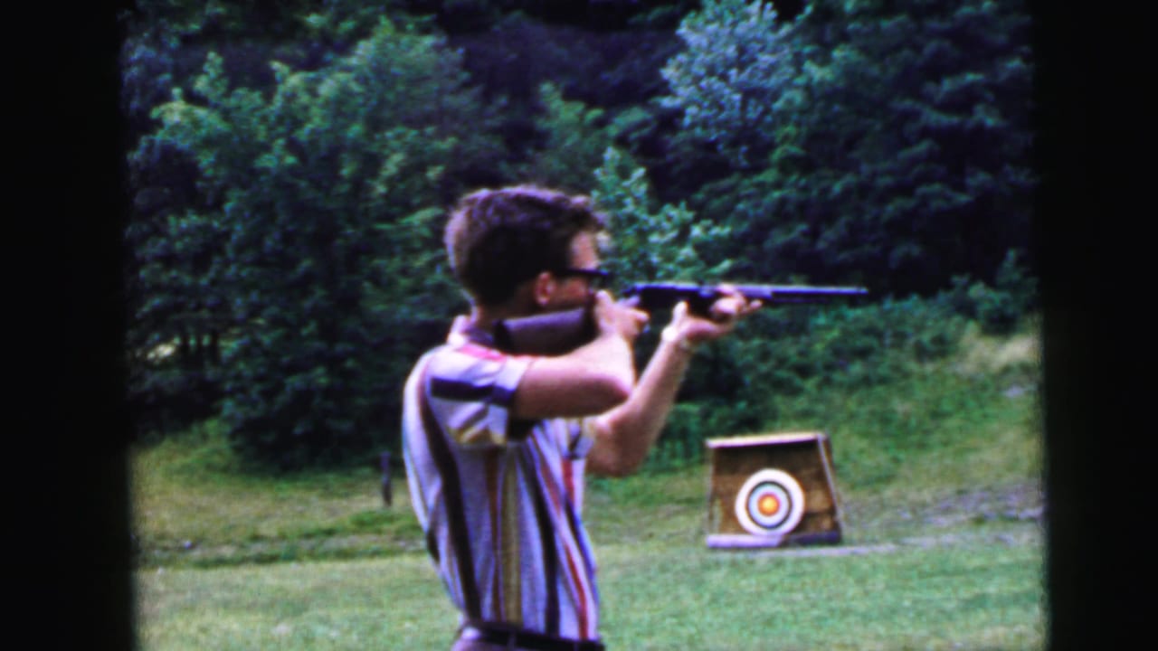 Young man in striped shirt and glasses aiming rifle to the right; circular bullseye target visible on stand in background.