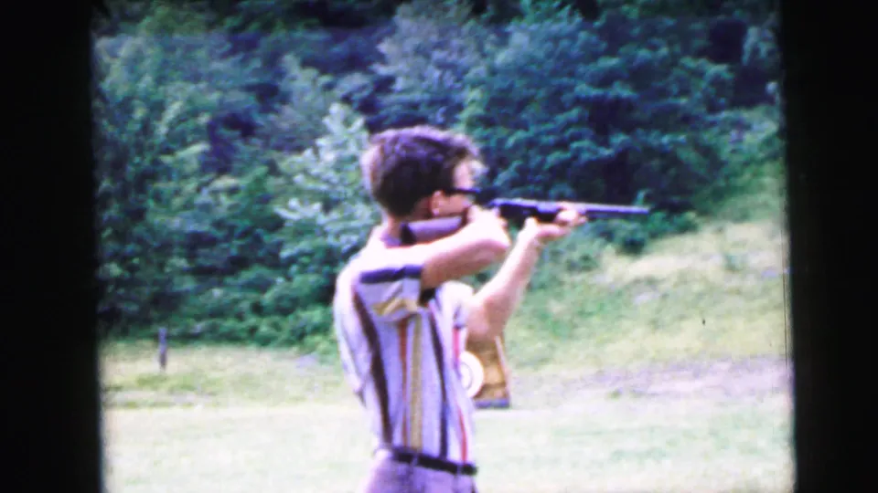 Three-quarter rear view of young man in striped shirt and glasses aiming rifle across a green field; partial bullseye target visible at lower right, 1964 Camden NJ 8mm home movie.