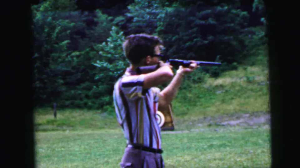 Young man in striped shirt and glasses aims rifle in profile at outdoor shooting range; dense deciduous tree line behind, partial target visible at lower right, 1964 8mm home movie.