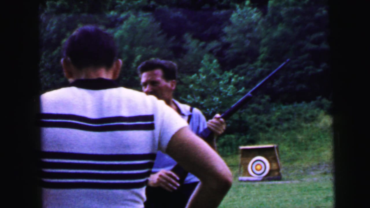 Two men at an outdoor shooting range — one in a striped shirt seen from behind, one in a light blue polo holding a rifle — with a bullseye target stand visible behind them.