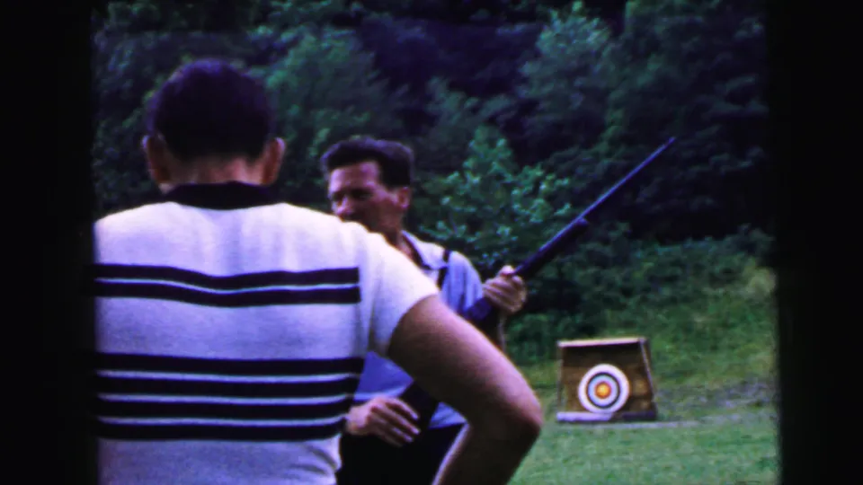 Two men exchange a rifle at an outdoor shooting range ca. 1964; one in a light blue polo faces the camera, the other in a striped shirt is seen from behind; circular bullseye target stand in background.