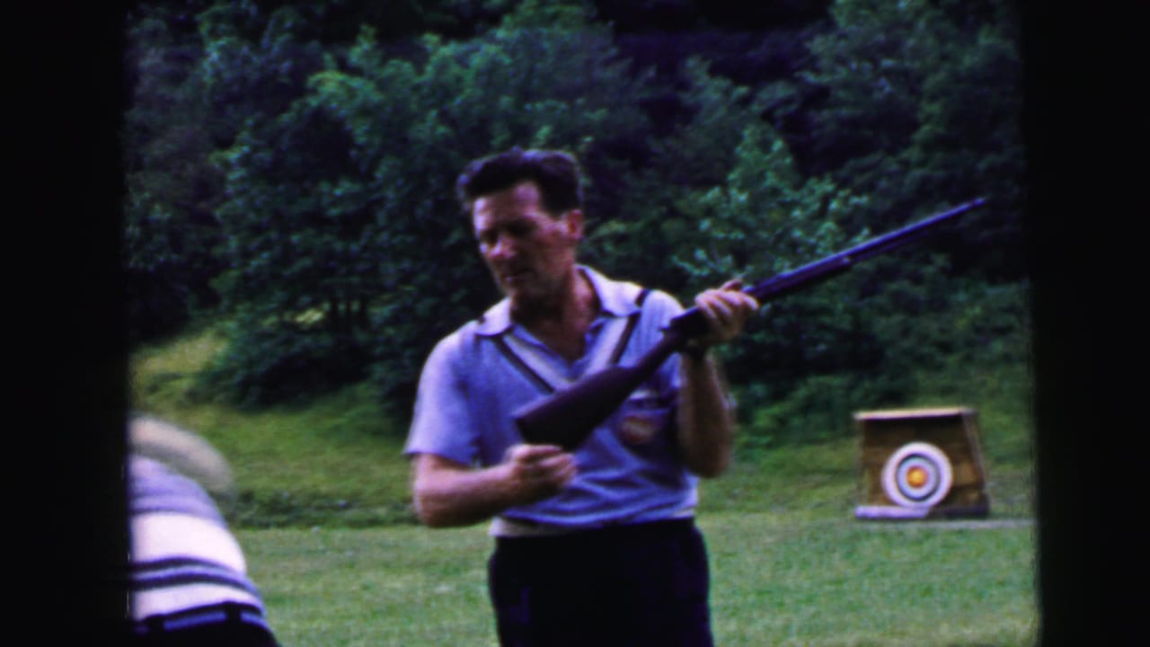 Smiling adult man in a light blue polo shirt holds a rifle at an outdoor shooting range ca. 1964; circular bullseye target on a wooden stand visible in background amid dense green trees.